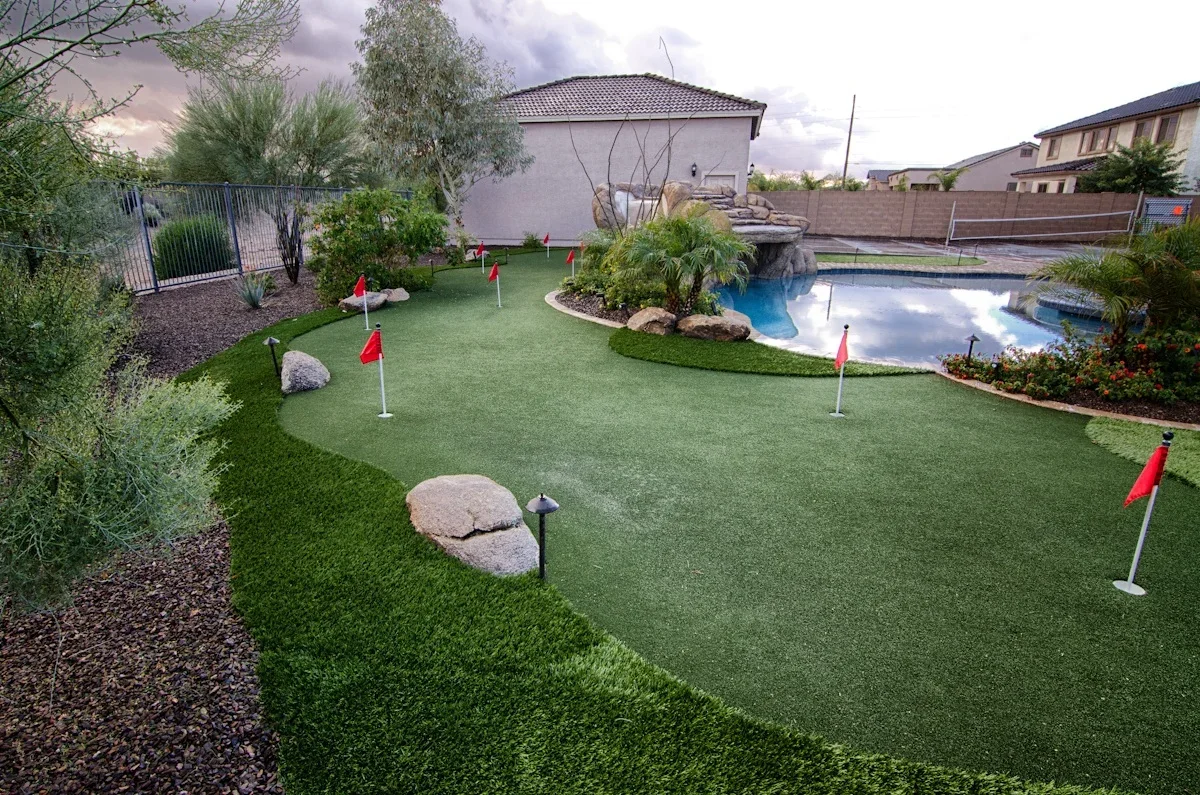 Synthetic putting green with red flags, boulders, and blue pool in residential backyard